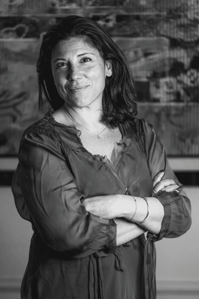 Portrait en noir et blanc D'Alexandra BERGHEIMER debout les bras croisés, devant un fond artistique dans son cabinet.