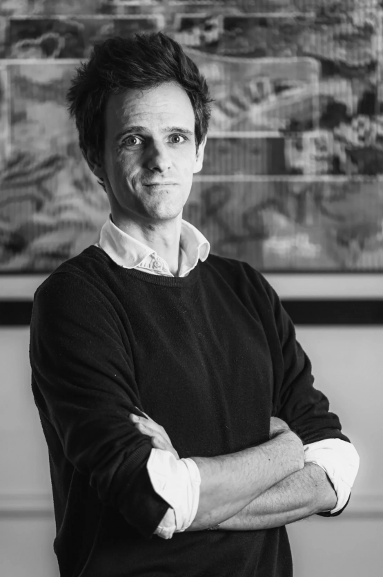 Portrait en noir et blanc d’un avocat du cabinet BERGHEIMER debout, les bras croisés, vêtu d’une chemise claire sous un pull sombre, devant un fond artistique